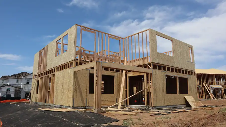 A two-story wooden house frame is under construction on a clear, sunny day.